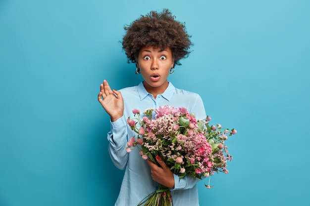 ショックを受けた巻き毛のアフロアメリカンの女性は 見知らぬ人から花束を受け取り 予期せぬ配達を受けたときにバグのある目を凝視し スタイリッシュな青い シャツを着て 屋内に立っています 花のコンセプト 無料の写真