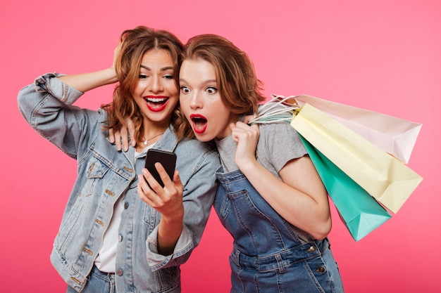 Shocked two women friends holding shopping bags using mobile phone. Free Photo