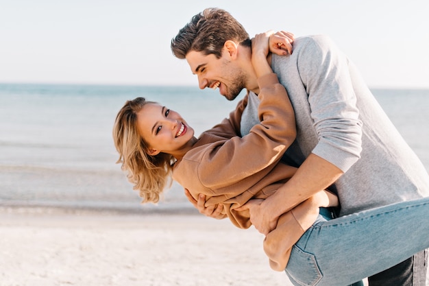 Short-haired blonde lady embracing husband in the beach. outdoor portrait of good-humoured man dancing with girlfriend near ocean. Free Photo