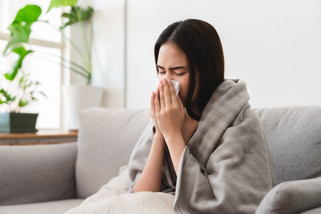 Sick asian young woman sneezing into tissue paper while covered with a blanket and sitting on sofa in the living room Premium Photo