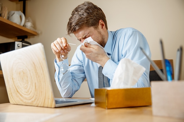 Sick man with handkerchief sneezing blowing nose while working in office, businessman caught cold, seasonal flu. Free Photo