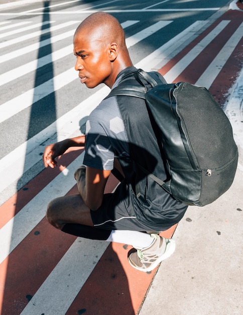 Side view of an african young healthy man with his backpack crouching on road in city Free Photo