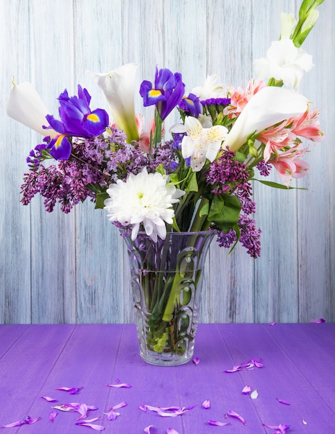 暗い紫色のアイリスライラックホワイトグラジオラスと灰色の木製の背景に紫の表面にガラスの花瓶にピンクのアルストロメリアの花と白い色のカラーリリーの花束の側面図 無料の写真