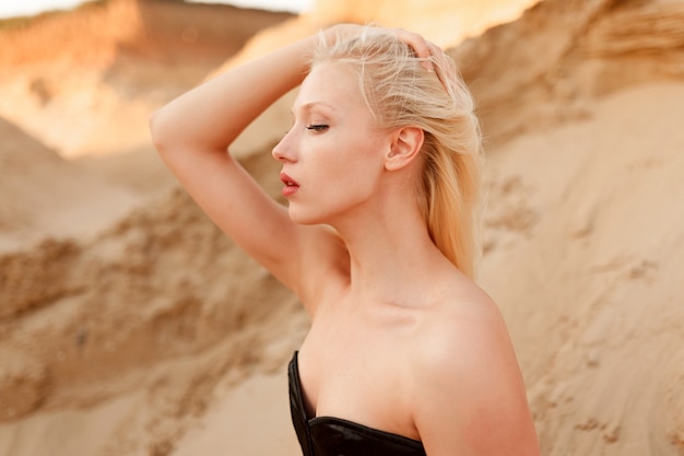 砂漠の砂の上に座って 彼女の髪に触れて 黒のセクシーなボディスーツで ブロンドの髪と化粧をした白人女性モデルの側面図 プレミアム写真
