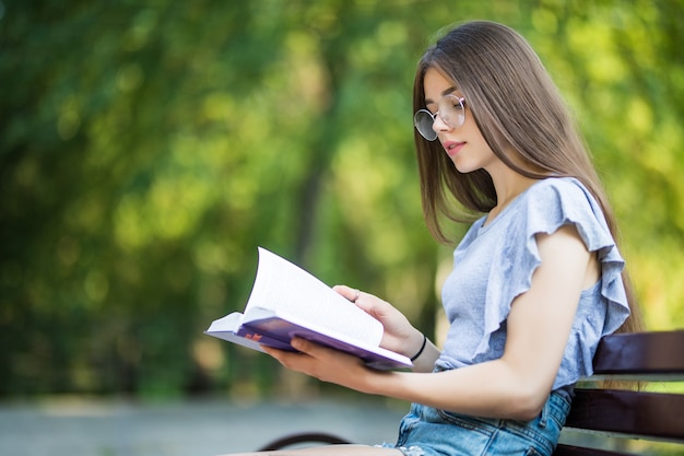 ベンチに座って公園で本を読んで眼鏡をかけて満足しているブルネットの女性の側面図 無料の写真