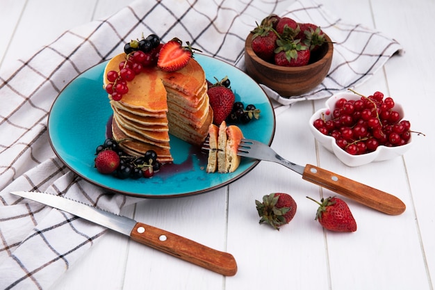 白の市松模様のタオルの上皿にフォークとナイフで黒と赤スグリのイチゴとサイドビューのパンケーキ 無料の写真