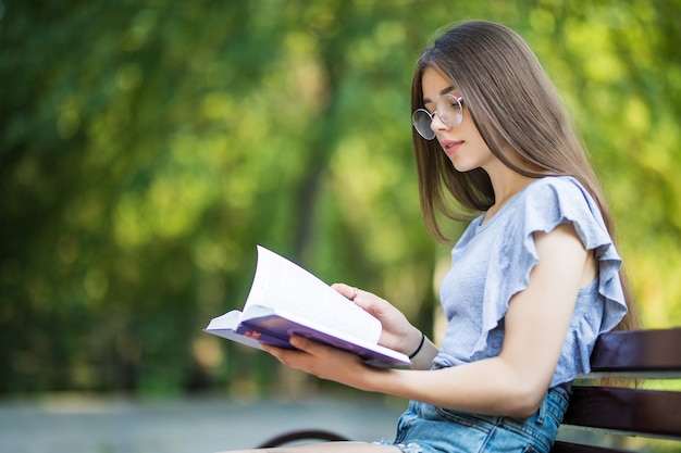 Side view of pleased brunette woman in eyeglasses sitting on bench and reading book in park Free Photo