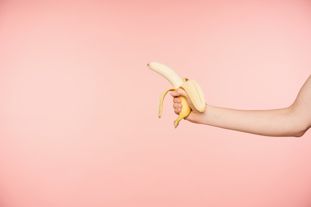 Side view of raised well-groomed female's hand with nude manicure holding peeled banana while posing over pink background, going to have healthy snack Free Photo