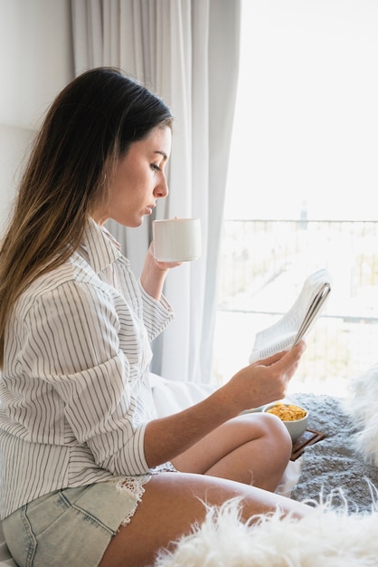 Side view of a woman sitting on bed drinking the coffee ...