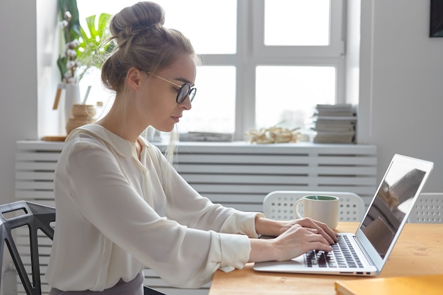 Sideways shot of serious fashionable young european businesswoman wearing stylish white blouse and round eyeglasses keyboarding on generic electronic device, checking email, writing business letter Free Photo
