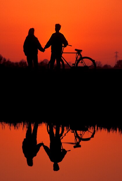 Silhouette of romantic couple with bicycle standing next to lake at sunset Premium Photo