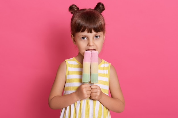 スリムな白人の女の子の子供は彼女の幸せな目で2つの大きなアイスクリームのルックスを保持し 面白い結び目を持っている ピンクの壁に孤立したポーズ 女性の 子供がおいしいアイスクリームをかむ 無料の写真