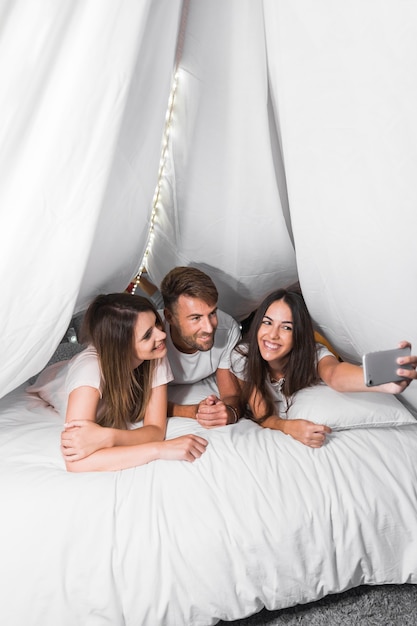 Free Photo Smiling Friends Lying On Bed Taking Selfie From Cellphone 