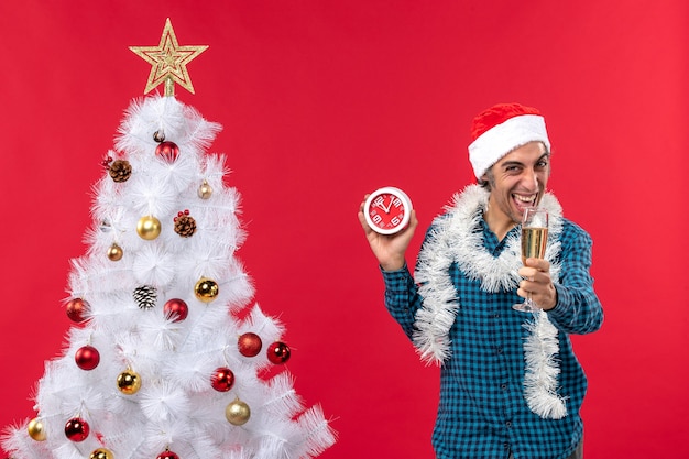 サンタクロースの帽子をかぶって面白い若い男を笑って ワインのグラスを上げて 赤いクリスマスツリーの近くに立っている時計を保持します 無料の写真