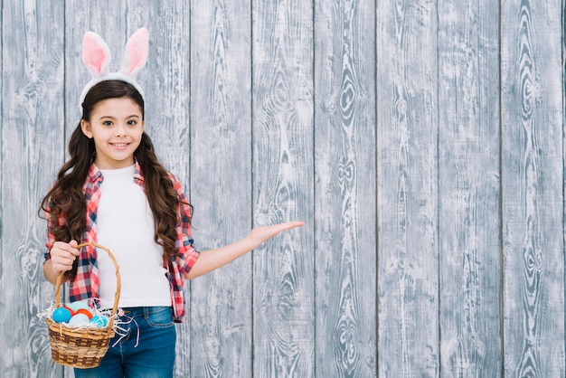 Smiling girl with bunny ears holding basket of easter eggs presenting against gray wooden desk Free Photo
