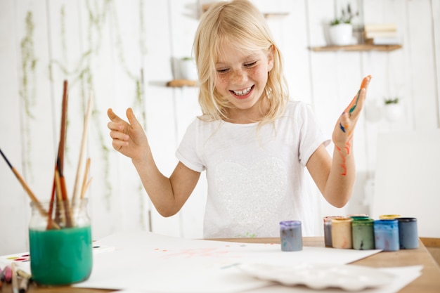 笑顔で 幸せで陽気なブロンドの女の子が彼女の歯を見せて 絵を描きながら楽しんでいます 女性のそばかすのある子供は さまざまな色の塗料で彼女の手を台無しにしました 無料の写真