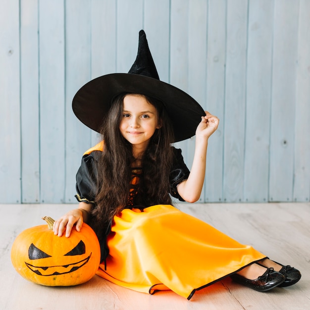 Free Photo | Smiling little witch sitting with pumpkin on floor