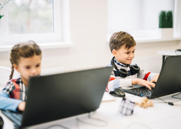 Smiling portrait of a boy using laptop sitting with a girl in the classroom Free Photo