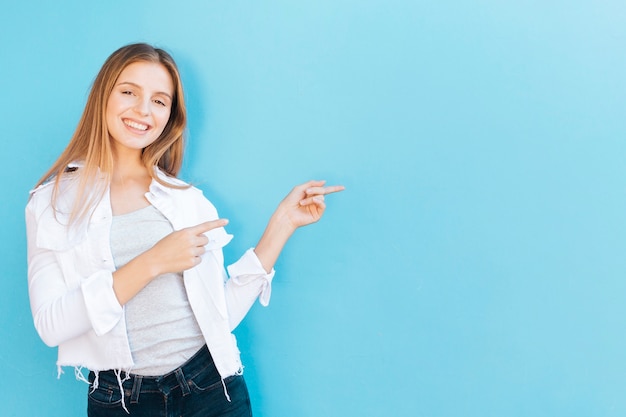 Smiling portrait of a young woman pointing her finger against blue background Free Photo