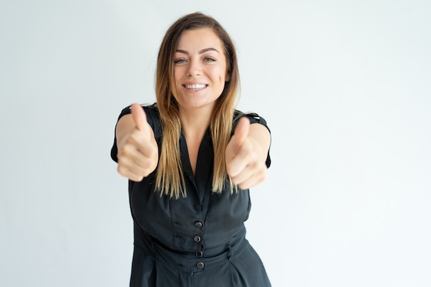 Smiling pretty young woman in black dress showing thumbs-up while expressing her approval Free Photo