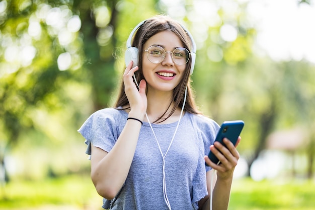 携帯電話を持って 公園を歩いて イヤホンで音楽を聴いて バックパックで若い女の子の学生を笑顔 無料の写真