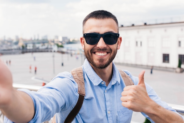 Smiling young man wearing sunglasses taking selfie and showing thumb up ...