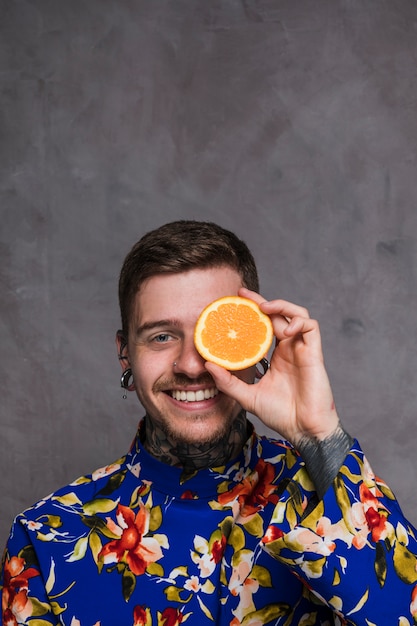 A smiling young man with piercing in the ears and nose holding slice of orange in front of his eyes against grey background Free Photo