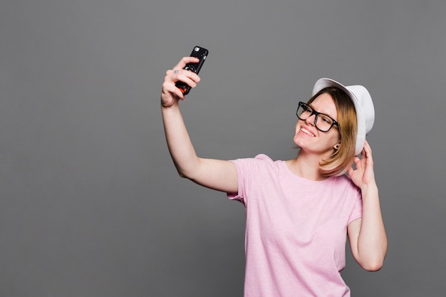 Smiling young woman wearing hat taking selfie on mobile phone against grey backdrop Free Photo