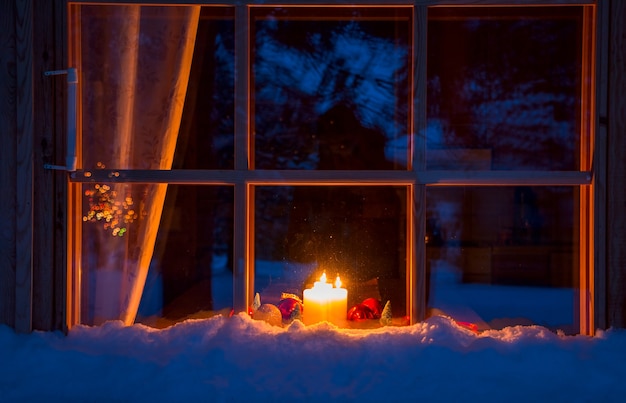 木造住宅の雪に覆われた夜の窓 窓辺にはクリスマスの飾りと燃えるろうそくがあります プレミアム写真