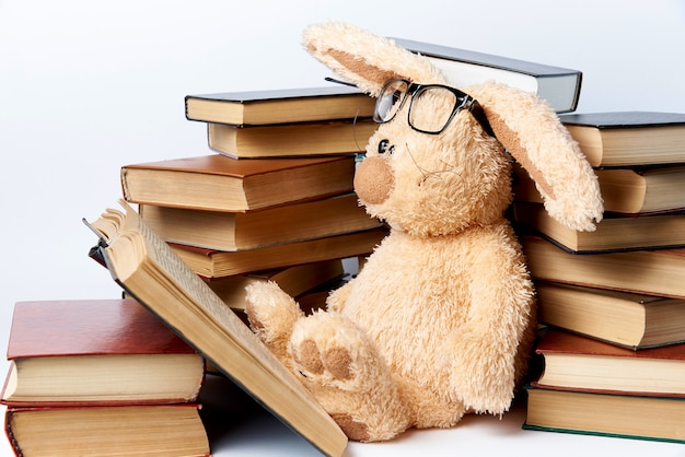 A soft toy rabbit in glasses sits in piles of books and reads a book. Premium Photo