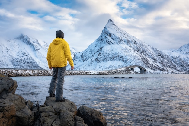 Sporty man in yellow jacket standing on the stone on seacoast against snowy mountains and cloudy sky