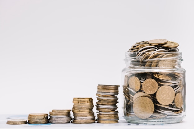 Stack of increasing coins with jar filled with coins against white backdrop Free Photo