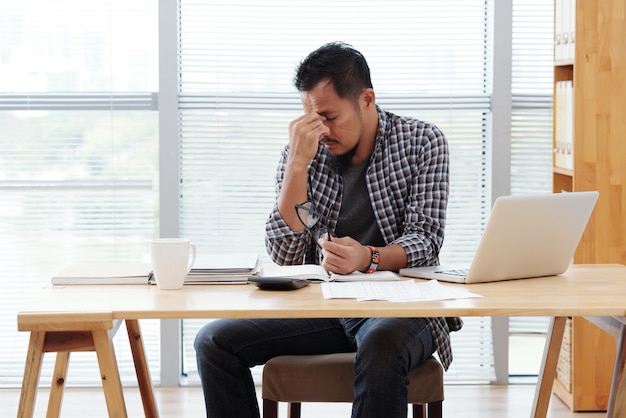 Stressed asian man sitting at table with laptop and documents and rubbing forehead Free Photo