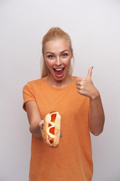 Free Photo Studio Photo Of Joyful Young Blue Eyed Blonde Lady With Casual Hairstyle Showing Hot Dog In Outstretched Hand And Raising Thumb Looking Happily At Camera And Smiling Widely