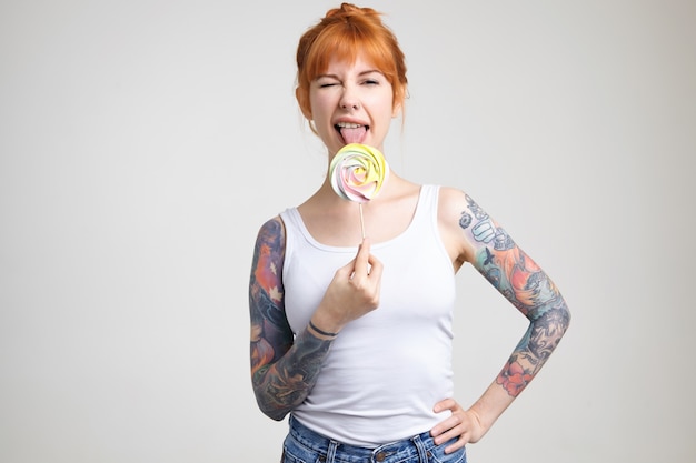 Free Photo Studio Photo Of Young Cheerful Tattooed Female With Foxy Hair Sticking Out Her Tongue While Licking Candy And Giving Wink At Camera Isolated Over White Background