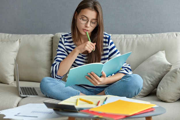 Studying online concept. serious young woman being busy with remote freelance project, sits at comfortable sofa, writes notes, holds textbook, use laptop computer at home with wireless internet Free Photo