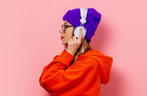 オレンジ色のパーカーとピンクの壁にヘッドフォンで紫の帽子のスタイルの女の子 プレミアム写真