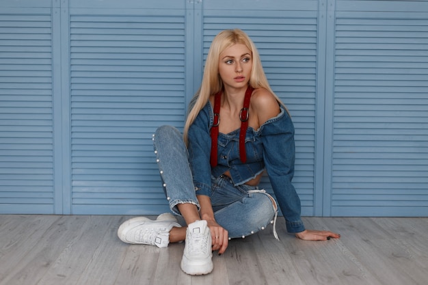 Premium Photo Stylish Blonde Woman In A Denim Shirt With Red Belts And Blue Jeans With White Shoes Sits Near Blue Wooden Wall