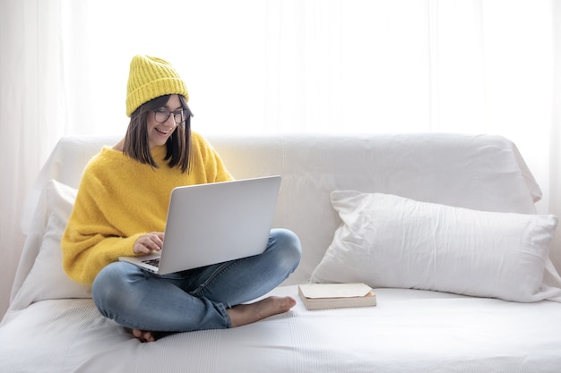 眼鏡と帽子のスタイリッシュな陽気なブルネットの少女は、自宅のソファに座って、ラップトップで動作します 無料写真