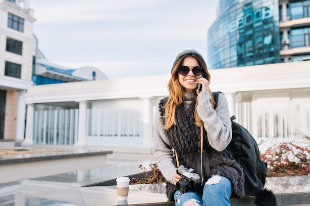 冬のウールのセーター サングラス ニット帽を身に着けているヨーロッパの近代的な市内中心部に座っているスタイリッシュな市うれしそうな若い女性 電話で話し バッグと一緒に旅行し カメラで 笑顔で テキストのための場所 無料の写真