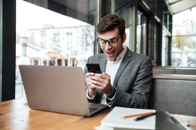Surprised happy businessman in eyeglasses sitting by the table in cafe with laptop computer and using smartphone Free Photo