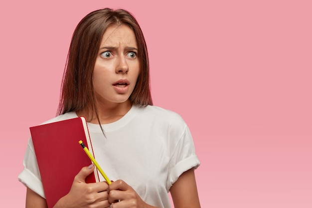 Surprised horrified young woman with dark straight hair, holds red textbook, pencil, feels puzzled of recieving many tasks Free Photo