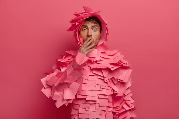 Surprised man covers mouth and stares with bugged eyes, afraids of something, hears astonishing news, gasps from shocking rumors, wears adhesive notes, isolated on pink wall. omg concept Free Photo