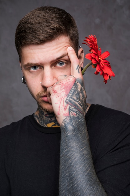 手に赤いガーベラの花を保持している彼の眉毛を上げる入れ墨の若い男 無料の写真