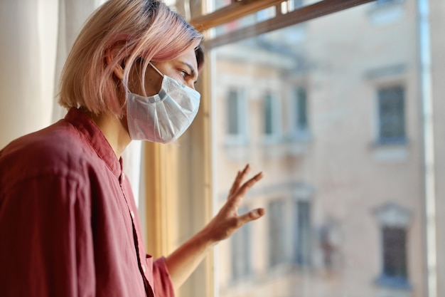 Teenage girl with pinkish hair standing in front of closed window with hand on glass, looking outside while staying at home during quarantine. coronavirus pandemic and social distancing concept Free Photo