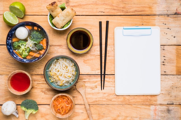 Thai soup; spring rolls; sauces and sprout beans with chopstick and white paper on clipboard over the wooden table Free Photo