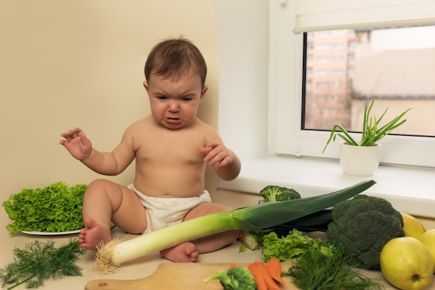 おむつの赤ちゃんは 新鮮な有機野菜や果物と一緒にキッチンテーブルに座っています 泣いて手を求めている子供 新鮮な有機野菜と果物で プレミアム写真