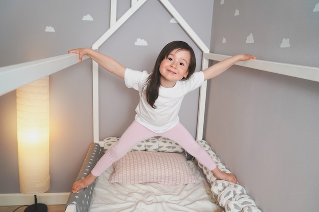 赤ちゃんは寝る前に彼のベッドでジャンプしています うれしそうな女の子がベッドにふける 白いtシャツとピンクのレギンスの幼児 プレミアム写真