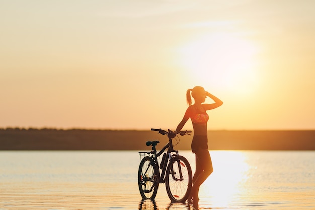 カラフルなスーツを着た強いブロンドの女性は 暖かい夏の日の日没時に水の中の自転車の近くに立っています フィットネスのコンセプト 無料の写真