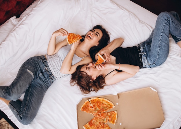 The stylish sisters eating slices of pizza and lying on the bed Free Photo The stylish sisters eating slices of pizza and lying on the bed Free Photo